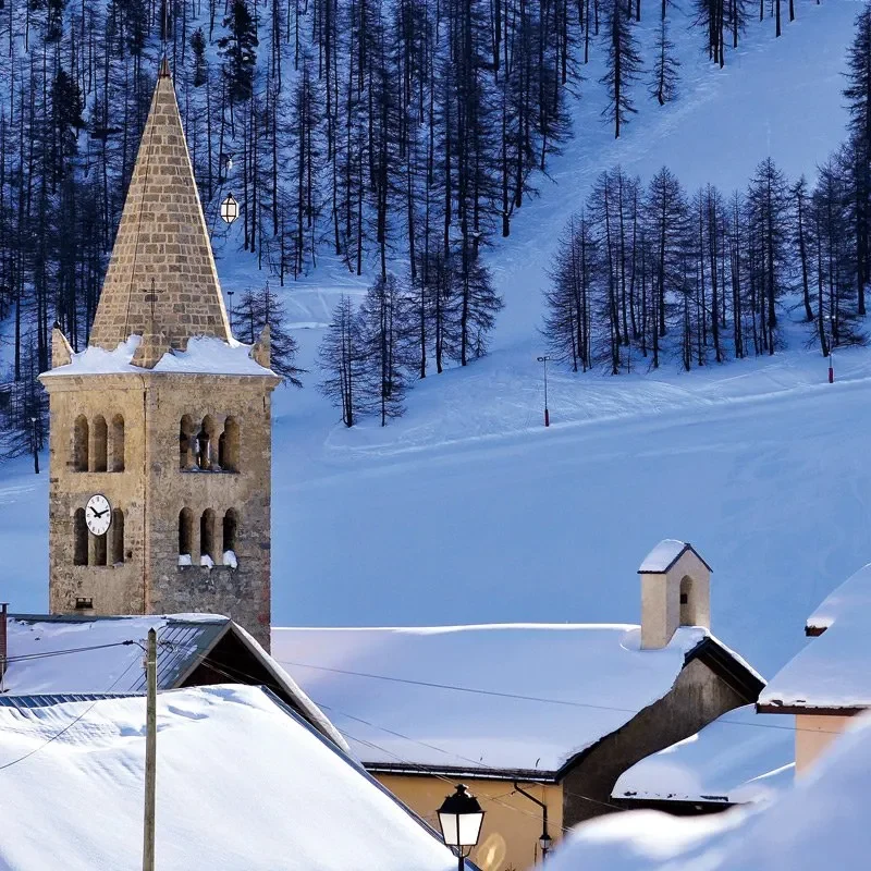 Église avec un clocher, entourée de montagnes enneigées et de maisons en bois.